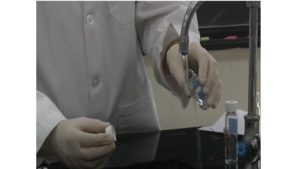 Person in lab coat rinsing a small vial under a faucet.